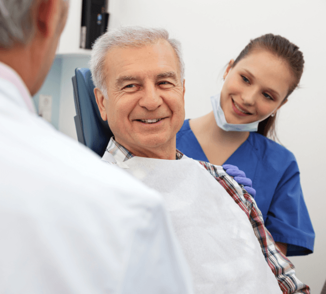 An older man listens closely as his dentist and dental assistant discuss the cost of dental implants