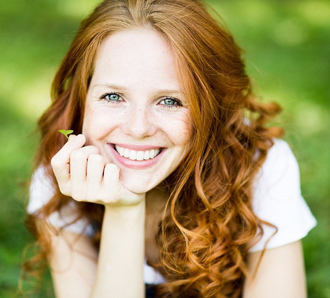 A young woman with red hair holding a small leaf and smiling while standing outside