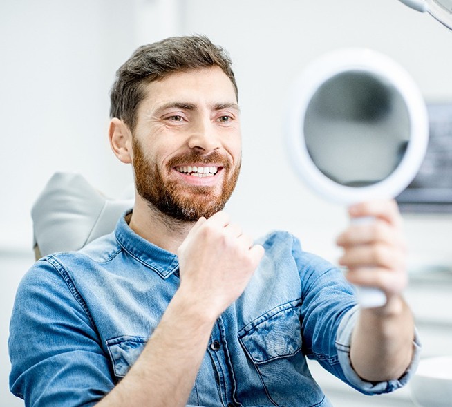 A man wearing a denim button-down shirt and holding a handheld mirror while smiling in the dentist’s office after seeing the result of his smile makeover